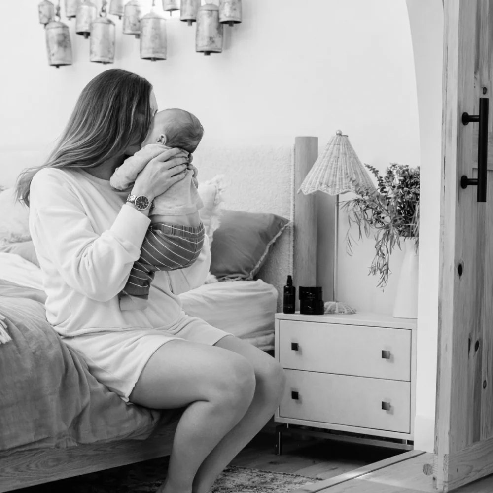 A mother holding and kissing her newborn in a sunlit bedroom